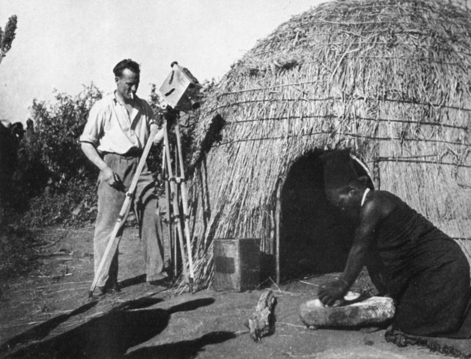 A man films a woman making food outside her straw hut